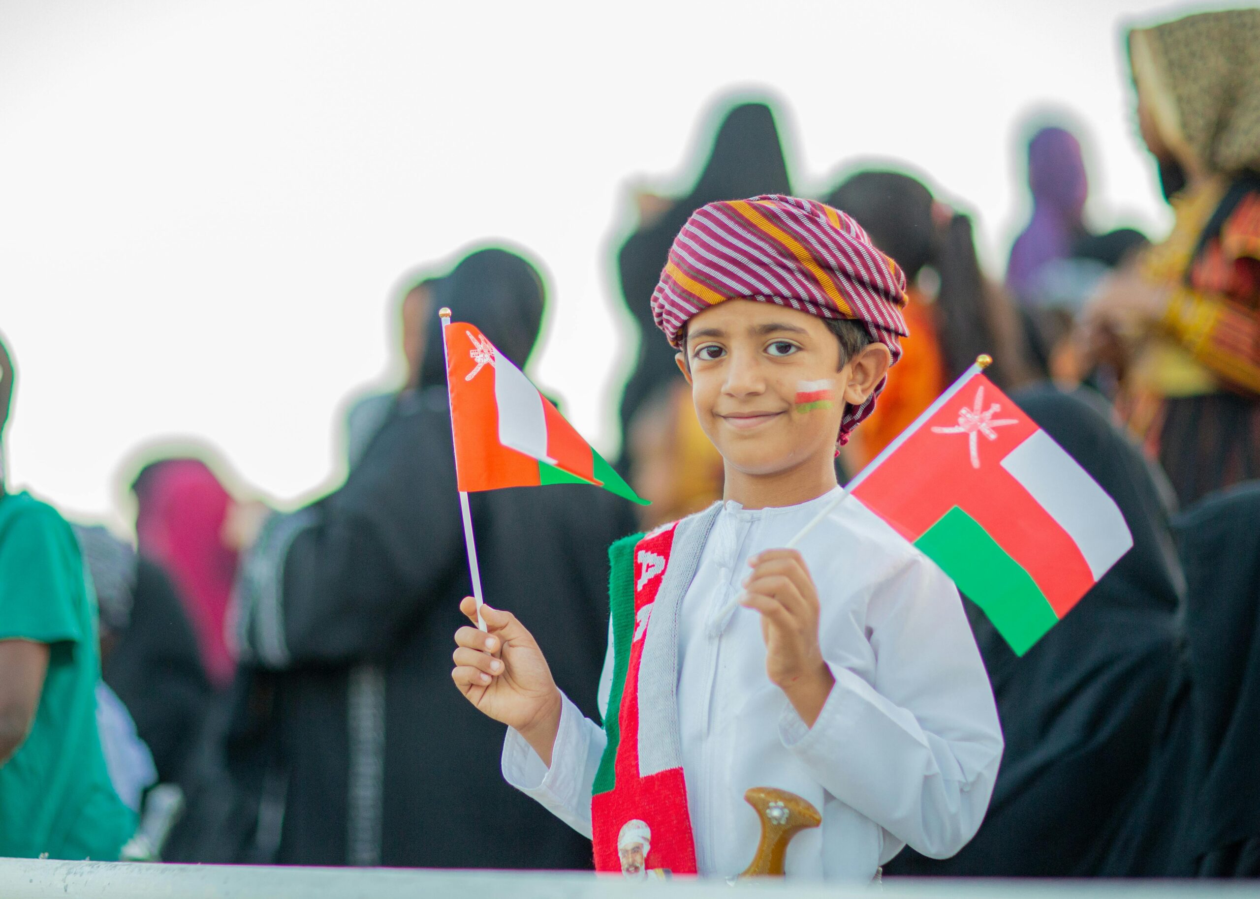 Young boy in traditional attire holding Oman flags during National Day celebration.